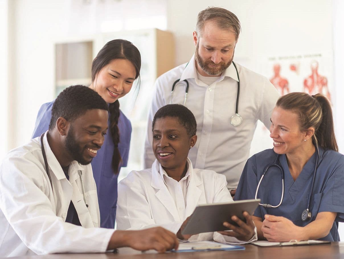 Five healthcare professionals in scrubs and white coats looking at a tablet together